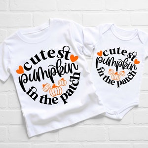 May include: Two white shirts with the text "cutest pumpkin in the patch" in black with orange pumpkins and red hearts. The larger shirt is a t-shirt and the smaller shirt is a onesie.