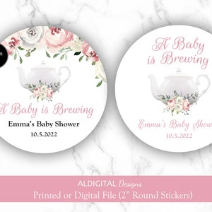 May include: Two designs for 2 inch round stickers for a baby shower. Design A features a teapot with pink and white flowers and the text "A Baby is Brewing Emma's Baby Shower 10.5.2022". Design B features a teapot with pink and white flowers and the text "A Baby is Brewing Emma's Baby Shower 10.5.2022".