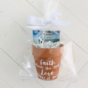 May include: A small terracotta pot with the text "Faith plants the seed Love makes it grow" in white lettering. The pot is filled with white shredded paper and a small blue packet of seeds.
