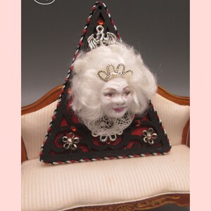 May include: A triangular wooden ornament with a black and red design. A white porcelain doll head with a crown is attached to the ornament. The ornament is sitting on a white couch.