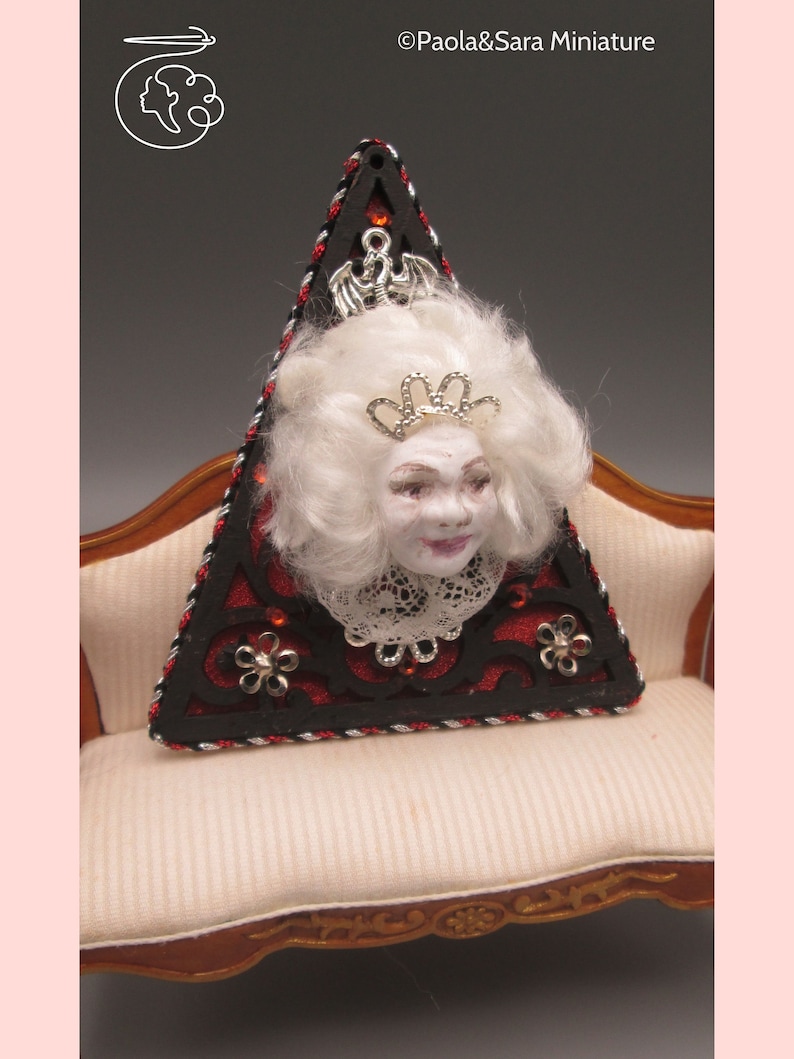 May include: A triangular wooden ornament with a black and red design. A white porcelain doll head with a crown is attached to the ornament. The ornament is sitting on a white couch.
