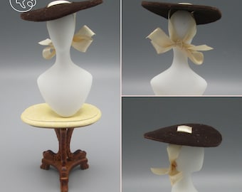 May include: A brown felt hat with a wide brim and a cream-colored satin ribbon tied in a bow. The hat is displayed on a white mannequin head.