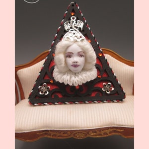 May include: A triangular wooden frame with a black and red design and a white porcelain doll head with a silver crown and a silver dragon charm. The frame is sitting on a white couch with a wooden frame.