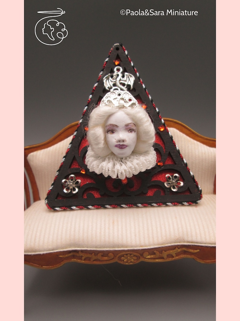 May include: A triangular wooden frame with a black and red design and a white porcelain doll head with a silver crown and a silver dragon charm. The frame is sitting on a white couch with a wooden frame.