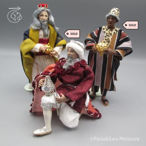 May include: Three miniature figurines representing the Three Wise Men. One figure kneels in a maroon robe holding a silver vessel, while two standing figures wear gold and striped robes, each holding a gold vessel. Two figures have SOLD tags.