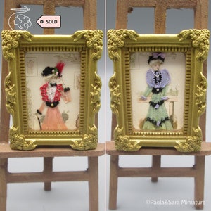 Könnte beinhalten: Zwei Miniatur-Bilder in goldenen Rahmen auf Staffeleien. Die Bilder zeigen Frauen in Vintage-Kleidung. Die Frau im linken Bild trägt ein rosa Kleid mit einer roten Jacke und einen schwarzen Hut mit einer roten Feder. Die Frau im rechten Bild trägt ein grünes Kleid mit einer lila Jacke und einen schwarzen Hut. Beide Bilder haben einen goldenen Rahmen.
