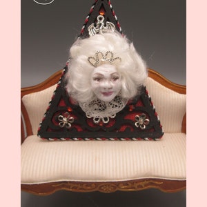 May include: A triangular wooden ornament with a black and red design. The ornament features a white doll's head with a crown and lace collar. The ornament is sitting on a white couch with gold trim.