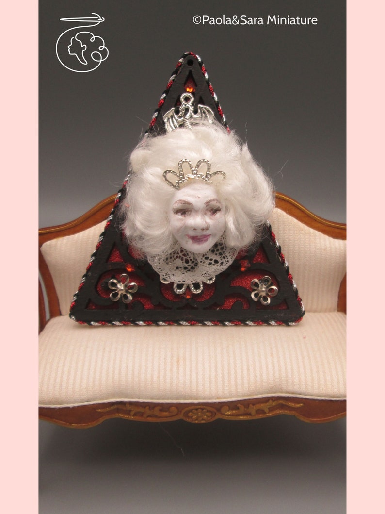 May include: A triangular wooden ornament with a black and red design. The ornament features a white doll's head with a crown and lace collar. The ornament is sitting on a white couch with gold trim.