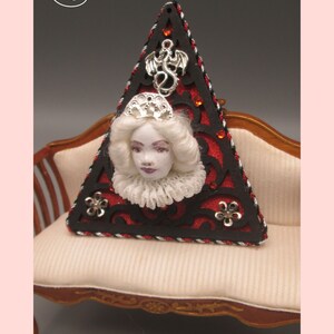 May include: A triangular red and black ornament with a silver dragon, a white porcelain doll head, and a silver crown. The ornament is sitting on a white couch with a wooden frame.