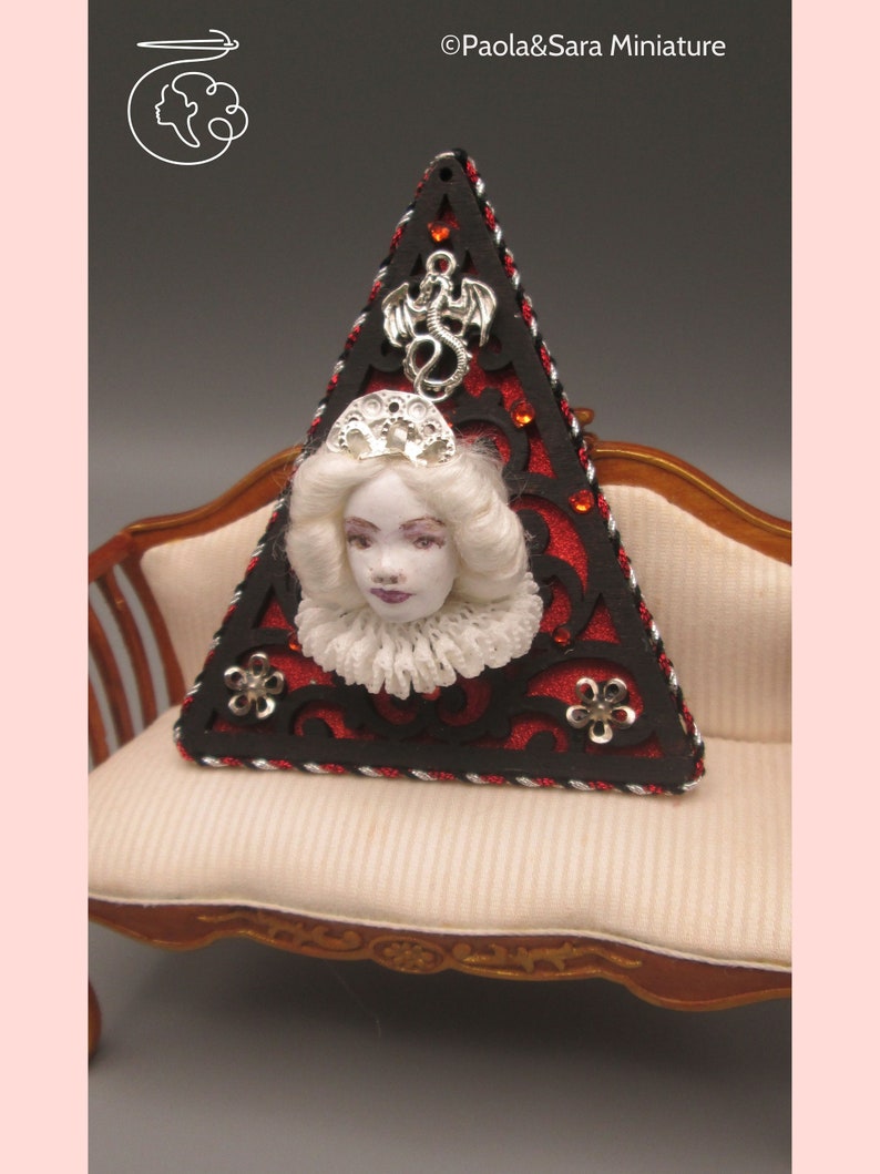 May include: A triangular red and black ornament with a silver dragon, a white porcelain doll head, and a silver crown. The ornament is sitting on a white couch with a wooden frame.