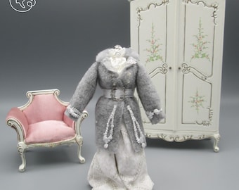 May include: A miniature doll wearing a grey coat with white trim and a white dress. The doll is standing in front of a white armoire with floral designs and a pink armchair.