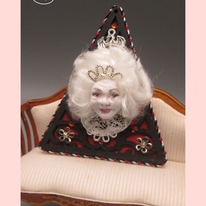 May include: A triangular shaped decorative piece with a red and black background. The piece features a white haired doll head with a silver crown and a lace collar. The piece is sitting on a white couch with gold trim.