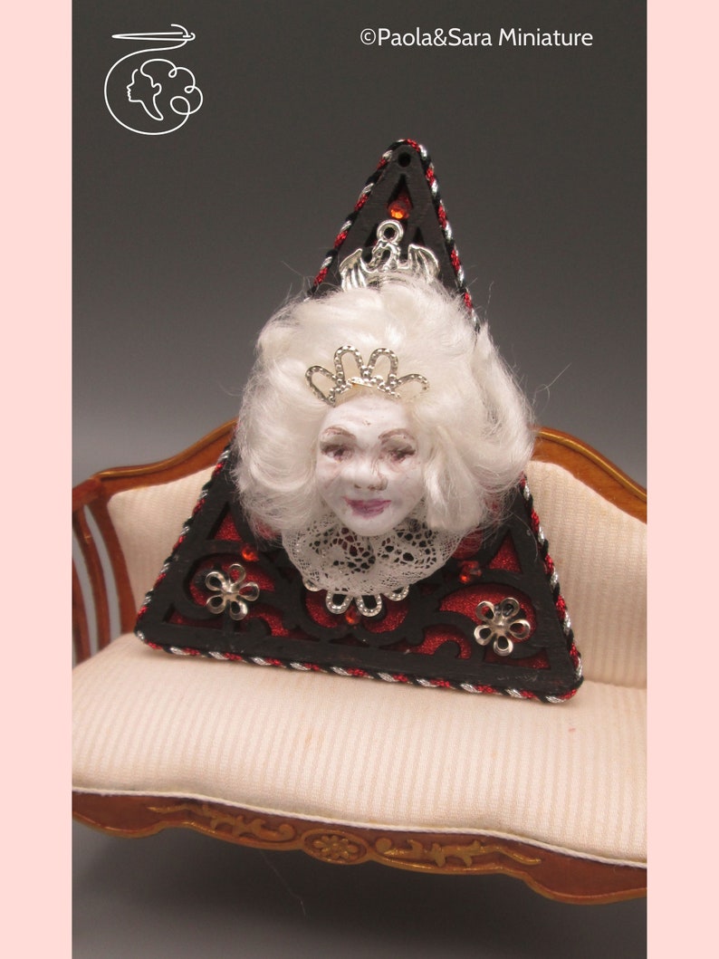 May include: A triangular shaped decorative piece with a red and black background. The piece features a white haired doll head with a silver crown and a lace collar. The piece is sitting on a white couch with gold trim.