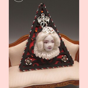 May include: A triangular, red and black, decorative piece with a white porcelain doll head and a silver dragon charm. The piece is sitting on a white, tufted sofa.