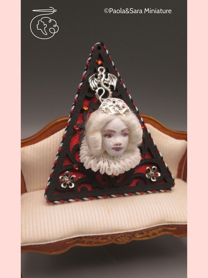 May include: A triangular, red and black, decorative piece with a white porcelain doll head and a silver dragon charm. The piece is sitting on a white, tufted sofa.