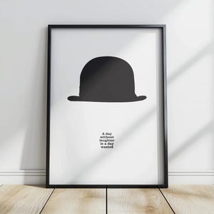 Può includere: Stampa incorniciata con una silhouette di bombetta nera su sfondo bianco. Il testo "A day without laughter is a day wasted" è incluso.