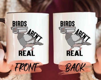 May include: White ceramic mug with a black handle and rim. The mug features a cartoon pigeon wearing a surveillance camera, with the text "BIRDS AREN'T REAL" in black. The front of the mug says "FRONT" in a pinkish-orange color.