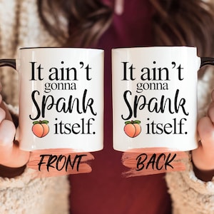 May include: White ceramic mug with a black rim and handle. The mug has a peach graphic and the text "It ain't gonna Spank itself." on the front and back.