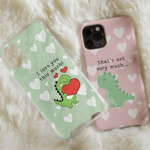 Puede incluir: Dos fundas para teléfono con un diseño de dinosaurio verde. Una funda es verde con corazones blancos y el texto "¡Te quiero tanto!" La otra funda es rosa con corazones blancos y el texto "Eso no es mucho..."