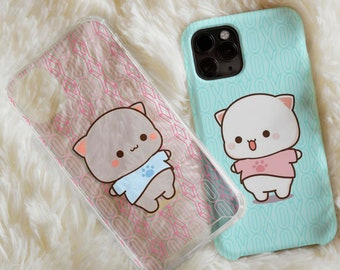 May include: Two clear phone cases featuring cute cartoon cat designs. One case has a pink and white pattern with a grey cat wearing a blue shirt. The other case has a turquoise and white pattern with a white cat wearing a pink shirt.