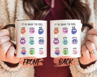 May include: White ceramic mug with a black handle. The mug has a watercolor design of different emotions with the text "It is okay to feel" on the front and back.