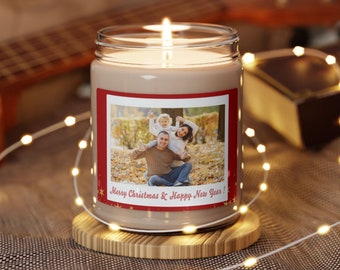 May include: A lit candle in a clear glass jar with a family photo label. The label has a red border and the text "Merry Christmas & Happy New Year!". The candle sits on a wooden coaster, with string lights and a guitar in the background.