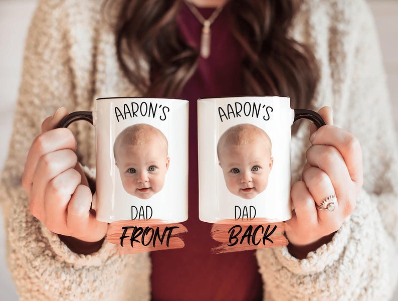 A person is holding two white mugs with black text and a photo of a baby's face on them.