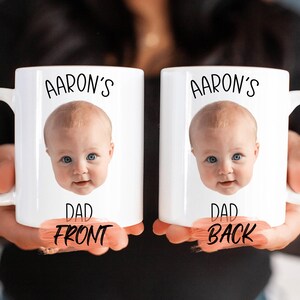 Two white mugs with a personalized photo of a baby's face printed on them, along with the text "AARON'S" and "DAD FRONT" printed below the image.