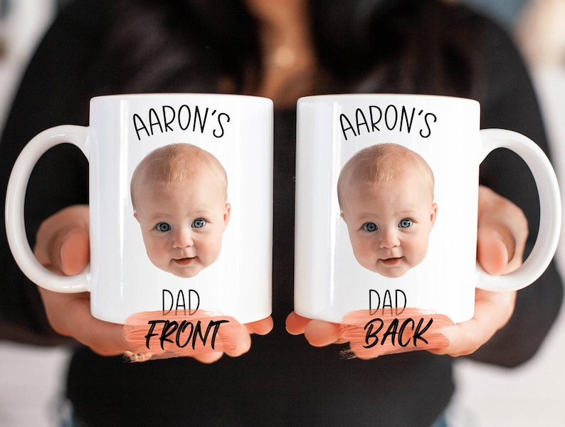 Two white mugs with a personalized photo of a baby's face printed on them, along with the text "AARON'S" and "DAD FRONT" printed below the image.