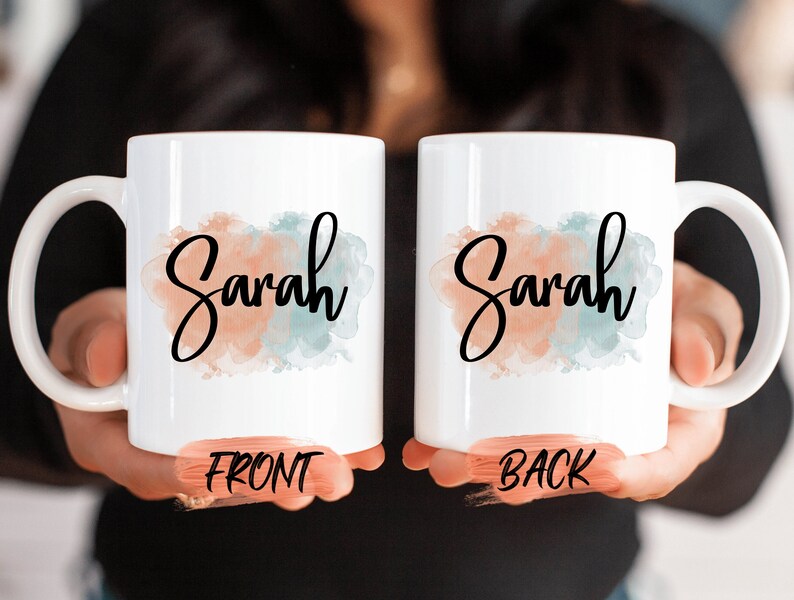 Op de afbeelding: Twee witte keramische mokken met een aquarelontwerp. De voorkant van de mok zegt "Sarah" in zwarte schrijfletters met een perzikkleurige en blauwe aquarel achtergrond. De achterkant van de mok zegt "Sarah" in zwarte schrijfletters met een blauwe en groene aquarel achtergrond.