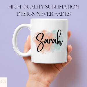 Op de afbeelding: Witte keramische mok met een aquarelontwerp in tinten perzik en blauw. De naam "Sarah" staat in zwarte cursieve letters op de mok.