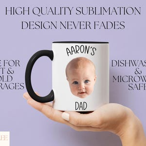 A person's hand holding a white mug with a black handle, featuring a photo of a baby's face.