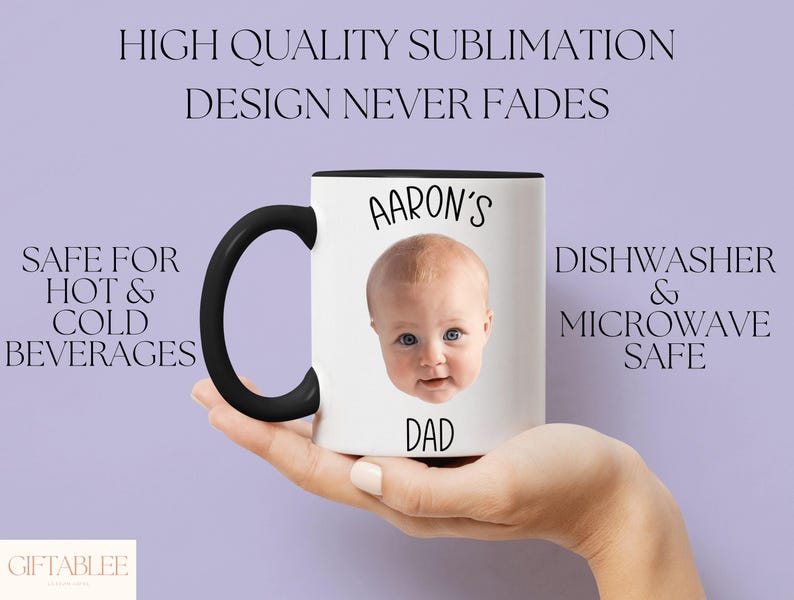 A person's hand holding a white mug with a black handle, featuring a photo of a baby's face.