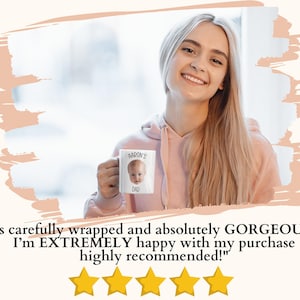 A smiling woman holding a mug with a photo of a baby on it, standing in front of a window.
