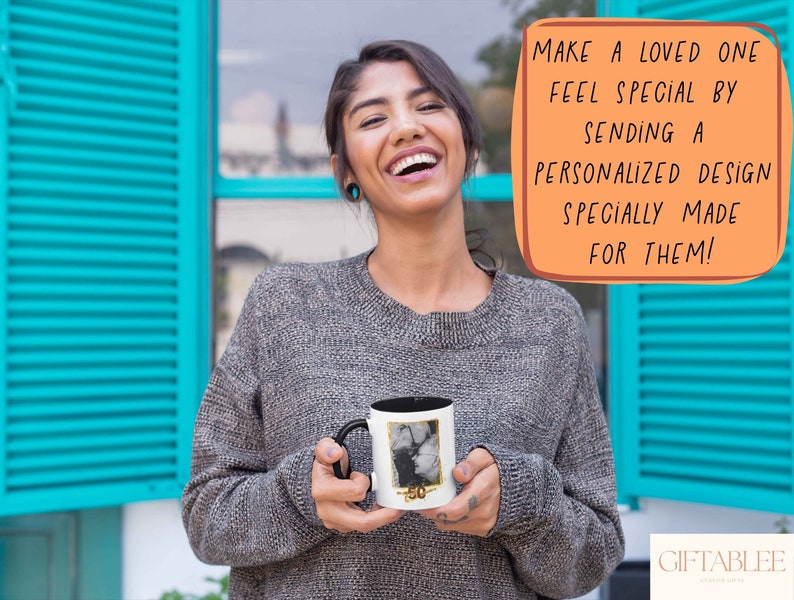 May include: A woman is holding a white mug with a black handle. The mug has a photo printed on it. The text on the mug says "50". The woman is smiling and looking at the camera. The background is a blue wall with white shutters.