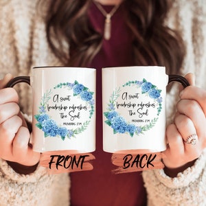 Puede incluir: Taza de cerámica blanca con asa negra. La taza tiene un diseño floral azul y el texto "Una dulce amistad renueva el alma" con la referencia bíblica "Proverbios 27:9".