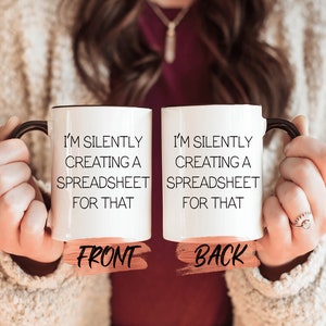 Puede incluir: Taza de cerámica blanca con asa negra. La taza tiene texto negro que dice "Estoy creando silenciosamente una hoja de cálculo para eso". La taza se muestra desde el frente y la parte posterior.