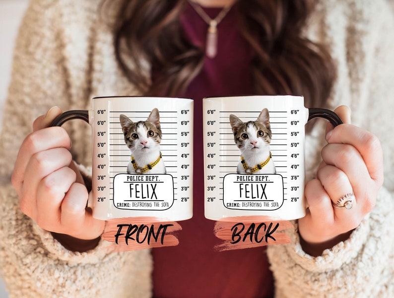 May include: White ceramic mug with a black handle. The mug features a black and white photo of a cat with a gold chain necklace. The mug has a white background with black lines and text that reads "Police Dept. Felix Crime: Destroying the Sofa".