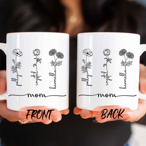 May include: White ceramic mug featuring a floral design with the names "pally", "crystal", and "karmella" in a script font. The word "mom" is written below the names. The mug has a curved handle and is held by a person.