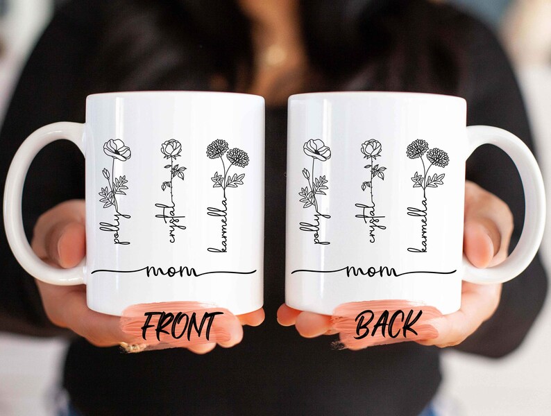 May include: White ceramic mug featuring a floral design with the names "pally", "crystal", and "karmella" in a script font. The word "mom" is written below the names. The mug has a curved handle and is held by a person.