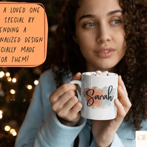 Op de afbeelding: Een witte keramische mok met een aquarelontwerp in tinten blauw, roze en oranje. De naam "Sarah" staat in zwarte schrijfletters op de mok. De mok is gevuld met marshmallows en wordt vastgehouden door een persoon.