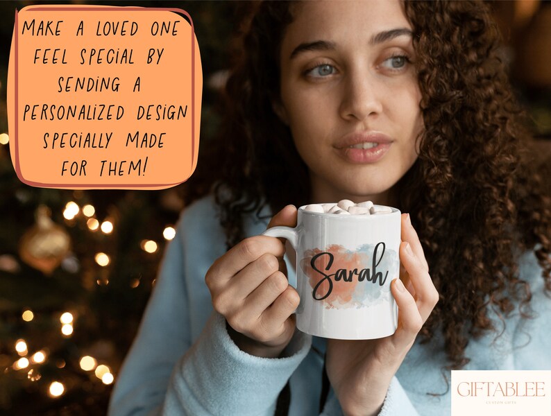 Op de afbeelding: Een witte keramische mok met een aquarelontwerp in tinten blauw, roze en oranje. De naam "Sarah" staat in zwarte schrijfletters op de mok. De mok is gevuld met marshmallows en wordt vastgehouden door een persoon.