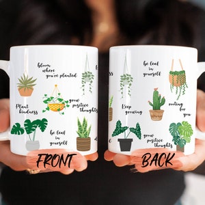 May include: White ceramic mug with a plant theme. The front of the mug features illustrations of various plants with text that reads "Plant kindness", "Bloom where you're planted", "Be leaf in yourself", and "Grow positive thoughts". The back of the mug features similar illustrations and text that reads "Keep growing", "Be leaf in yourself", "Rooting for you", and "Grow positive thoughts".