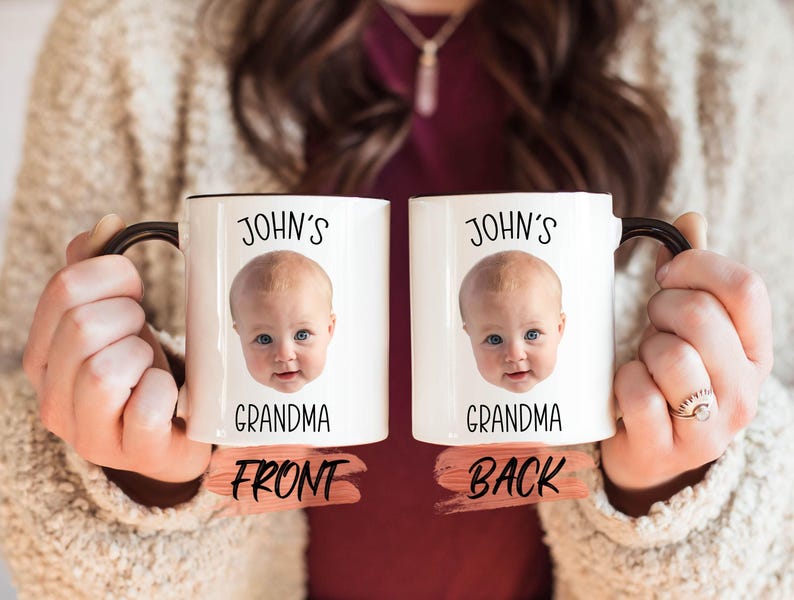 A person is holding two white mugs with black text and a photo of a baby's face on them.