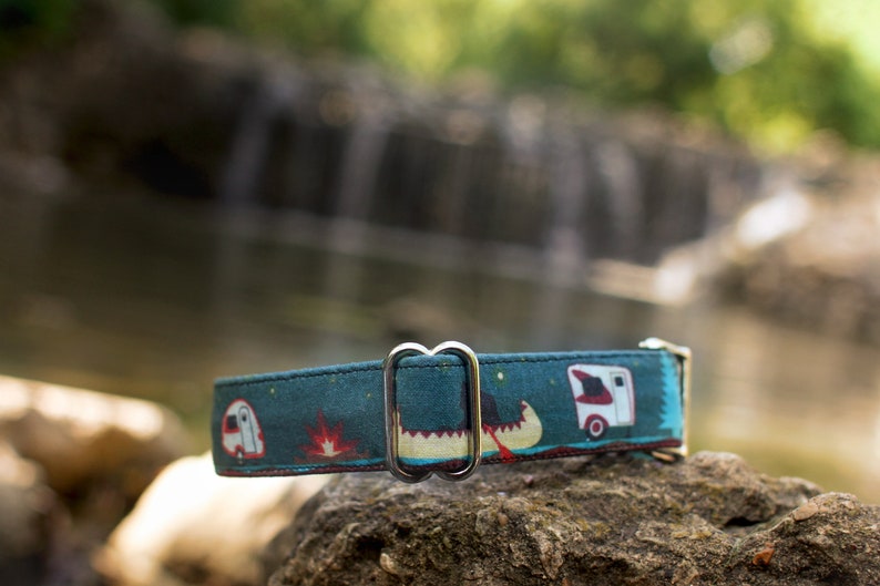 K&ouml;nnte beinhalten: Ein blaues Hundehalsband mit einem Camping-Motiv, das einen Wohnwagen, ein Kanu und ein Lagerfeuer zeigt. Das Halsband hat eine silberne Metallschnalle.