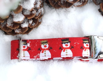 May include: Red dog collar with a snowman pattern and a silver buckle. The collar is lying on a snowy surface.