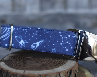 May include: A blue dog collar with a white space-themed print featuring stars, planets, and rockets. The collar has a silver buckle.