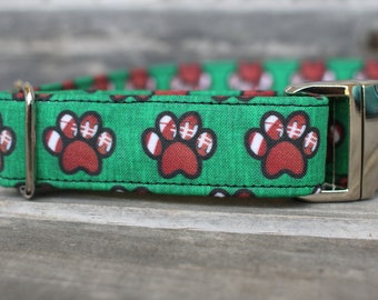 May include: Green dog collar with a red and white paw print pattern. The collar has a silver buckle.