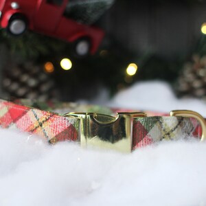 May include: A red and green plaid dog collar with a gold buckle. The collar is lying on a white fluffy surface.
