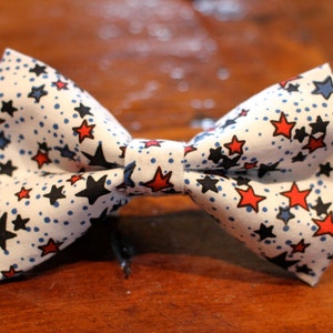 May include: A white bow tie with a red, white, and blue star pattern. The bow tie is made of fabric and has a classic bow tie shape.
